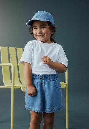 Smiling young girl wearing a white t-shirt, denim shorts, and a blue cap, standing indoors near a yellow metal chair against a gray background.