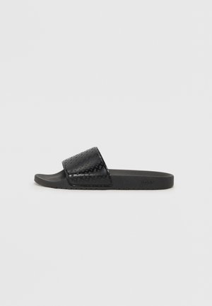 Schwarze Slip-On-Sandale mit strukturiertem breitem Riemen und flacher Sohle, Seitenansicht, minimalistisches Design auf einfarbig hellem Hintergrund.