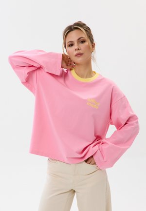 Vrouw in losse roze trui met gele kraag en tekst "offline mode", beige broek, poseren met een hand achter de nek en de andere op de heup.