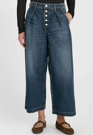 Femme porte un jean bleu taille haute à jambes larges avec des boutons visibles à l'avant, associé à des chaussures plates marron et un haut noir à manches longues.