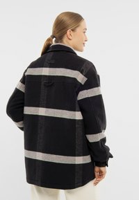 Černá a šedá kostkovaná flanelová bunda s zelenými akcenty, volný střih, špičatý límec a nastavitelné manžety; obsahuje zadní yoke a logo štítek.