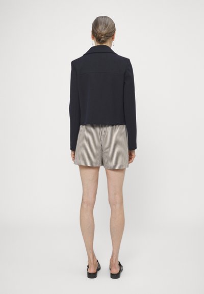 Veste cropped navy avec un col structuré, assortie à un short beige à rayures. Le tissu semble lisse, avec une coupe ajustée.