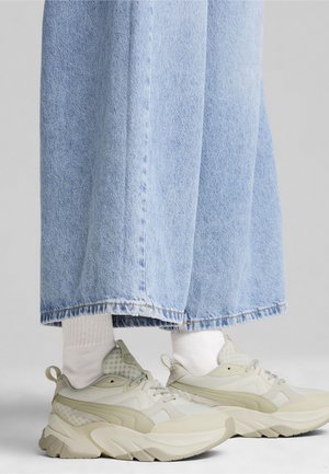 Pieds portant des baskets beige épaisses avec des chaussettes blanches et un jean large bleu clair.