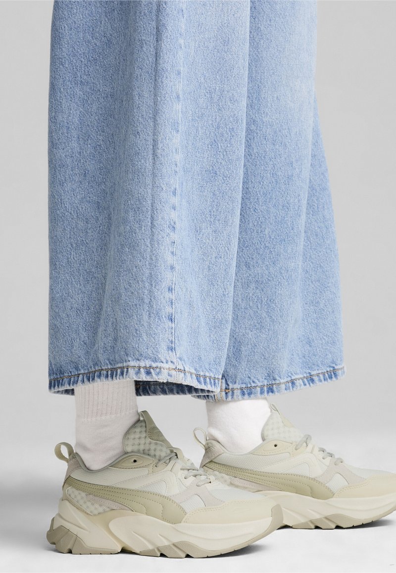Pieds portant des baskets beige épaisses avec des chaussettes blanches et un jean large bleu clair.