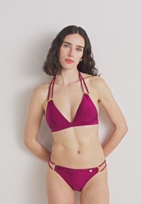 Magenta színű bikini szett, amely háromszögalakú, vékony pántos toppal és arany díszítéssel rendelkezik, valamint egy hozzáillő alacsony derekú alsó résszel és minimális textúrával.