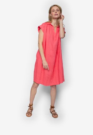 Vrouw die tegen een witte achtergrond staat, draagt een loszittende koraalkleurige jurk en bruine veter-sandalen, en raakt haar haar aan met één hand.