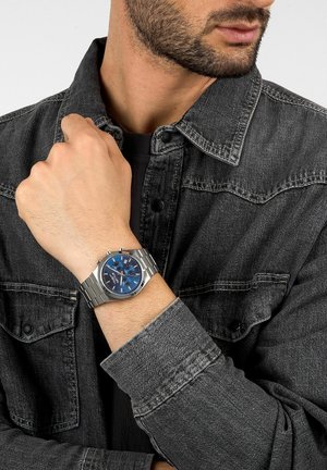 Orologio in acciaio inossidabile con quadrante blu e accenti argento, indossato al polso, posizionato su una giacca di jeans grigia. Quadrante circolare con funzioni cronografo.