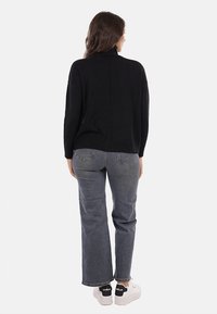 Donna in piedi di spalle, indossando un maglione dolcevita nero, jeans grigi e sneakers bianche con dettagli neri sul tallone.