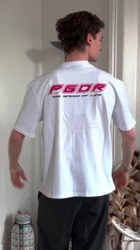 Weißes T-Shirt mit kurzen Ärmeln, das einen großen pinken "PGDR" Schriftzug und einen kleineren "THE SPEED OF LIFE" Schriftzug auf der Rückseite zeigt. Einfaches Design und Schnitt.