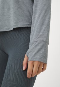 Harmaa pitkähihainen paita, valmistettu pehmeästä kankaasta, yhdistetty tummanharmaisiin leggingseihin, joissa on kohokuvioitu siksak-kuviointi ja peukaloreiät hihoissa.