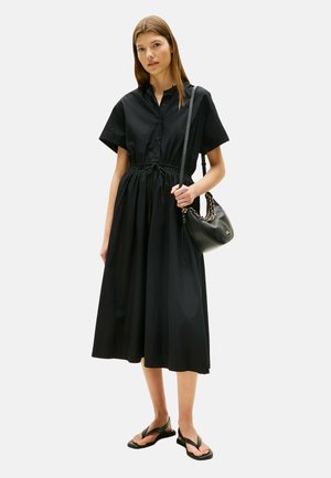Donna in piedi con un vestito midi nero con bottoni, vita regolabile con cordoncino, sandali neri e una borsa nera con tracolla a catena.