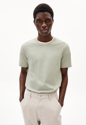 T-shirt vert clair à manches courtes en coton, col rond, porté avec un pantalon beige doté de poches latérales et d'un détail de couture.
