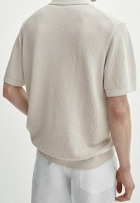 Beige neulepikeepaita, jossa on lyhyet hihat, teksturoitu kangas sekä resorit kauluksessa ja helmassa. Takana on rento istuvuus ja hienovarainen kuvio.