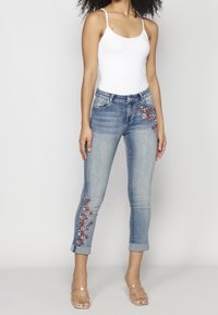 Hellblaue Jeans aus Denim mit roter und weißer Blumenstickerei an den Seiten, mit schmalem Bein und umgekrempelten Bündchen. Transparente Sandalen getragen.