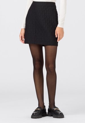 Woman wearing a black textured mini skirt, sheer black tights, black chunky loafers, and a long-sleeve white top against a plain background.