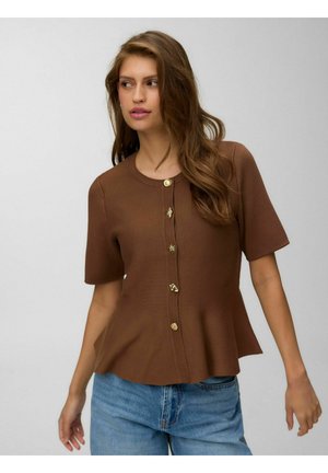 Ung kvinde med langt brunt hår iført en brun knapbluset bluse med korte ærmer og blå jeans, ser mod venstre med et neutralt udtryk.