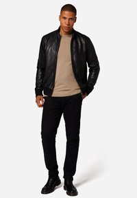 Svart läder bomberjacka, beige tröja, svarta byxor, svarta chelsea-boots. Modellen står upprätt med händerna i fickorna, neutral bakgrund.