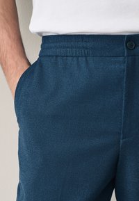 Close-up of a person wearing blue pants with a button and elastic waistband, hand resting in side pocket, and white shirt tucked in.