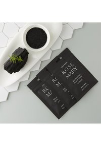Trois sachets noirs mats étiquetés "Rosemary Balancing Scalp Pack" avec du charbon. Un petit bol blanc contient de la poudre noire à côté d'un morceau de charbon.