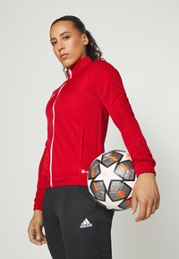 Rød sportsjakke med hvid lynlås, der har sidelommer, båret med sorte bukser med et hvidt Adidas-logo. Spilleren holder en fodbold.