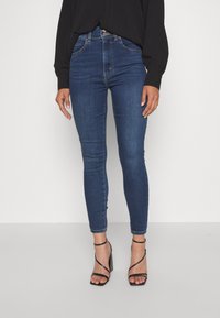 Högmidjade blå denim skinny jeans med en slät yta, femficksdesign och knappstängning, i kombination med svarta klacksandaler.