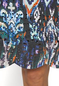 Tissu à motifs colorés avec des formes florales bleues, orange, blanches et noires et des détails découpés en cercles au-dessus d'une jambe brune.