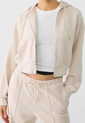 Gros plan d'une personne portant un sweat à capuche beige zippé, un pantalon beige assorti à cordon, et un crop top blanc côtelé sur un fond uni.