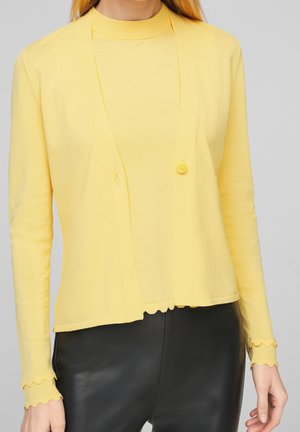 Kobieta ubrana w żółty kardigan z długimi rękawami z ząbkowanymi krawędziami, nałożony na dopasowany żółty top i czarne skórzane spodnie.