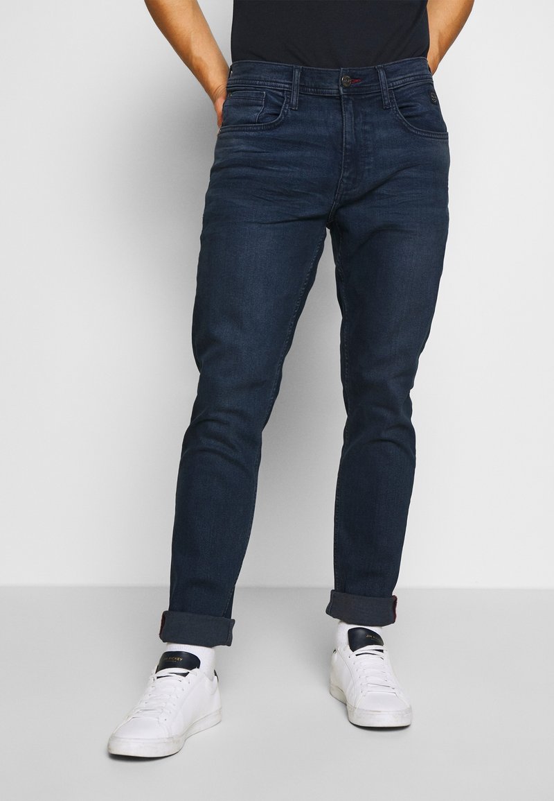 Homme portant un jean slim bleu foncé retroussé et des baskets blanches, debout devant un fond clair uni.