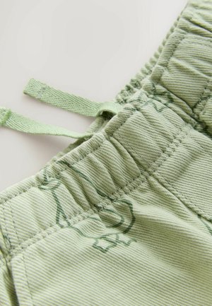 Közelkép halványzöld elasztikus derékpántól, húzózsinórokkal, texturált szövetnadrágon enyhe sötétzöld leveles mintával.