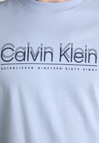 Šviesiai mėlynas medvilninis marškinėliai su užrašytu "Calvin Klein" ryškiu juodu šriftu ir plona linija apačioje, paprastas dizainas ir lygus paviršius.