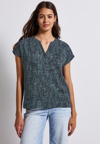 Green and black v-neck blouse with short sleeves, featuring a textured, abstract print. Paired with light blue jeans.