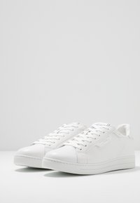 Zapatillas bajas blancas de cuero texturizado, con cordones planos, punta redondeada y un sutil detalle del logo en el lateral.