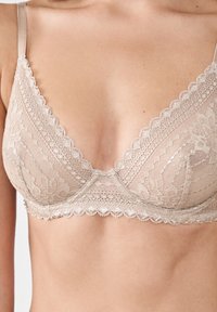Bralette de encaje en beige claro, con escote pronunciado, bordes festoneados y patrones florales que ofrecen una textura delicada. Tirantes ajustables.