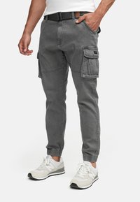 Homme portant un pantalon cargo gris avec des poignets élastiques, des baskets blanches et une ceinture noire, debout avec une main dans la poche.