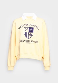 Gul sweatshirt med vit krage, mörkblå emblem med nycklar, lilja, ränder och ros, samt texten "Hollister California Laguna Hills Academy".