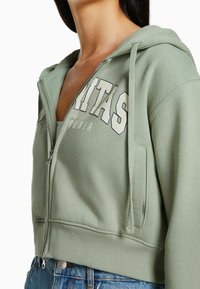 Mujer con una sudadera verde clara sin cerrar, con letras blancas, sobre una camiseta blanca y jeans azules, recortada por debajo del cuello.