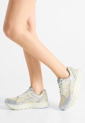 Ľahké športové topánky majú sieťovaný vrch v béžovej a striebornej farbe s reflexnými akcentmi, polstrovanou podrážkou a textúrovanými detailmi na päte.