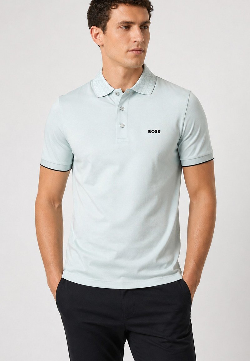 Homme portant un polo bleu clair à manches courtes avec le logo "BOSS" et un pantalon noir, debout les mains dans les poches devant un fond uni.