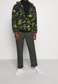 Uomo che indossa un giacca a fantasia verde e nera, un dolcevita nero, pantaloni verde oliva e sneakers bianche, in piedi contro un muro semplice.