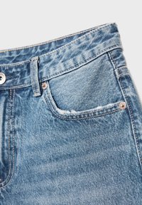 Detail světlých modrých džín s viditelným pasem, přední kapsou s kovovými nýty a zapínáním na knoflík na bílém pozadí.