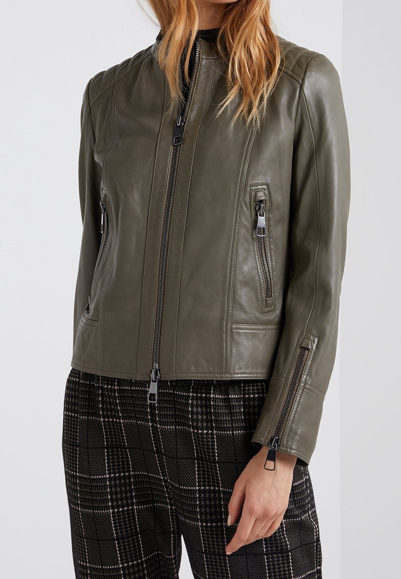 Oliivinvihreä nahkatakki, jossa on tiukka muotoilu. Varustettu vetoketjuilla etutaskuissa ja hihoissa, sileä pinta ja pystykohouma.