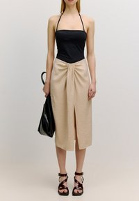 Zwarte haltertop, beige knielange rok met plooien en een voorknop, gecombineerd met bruine sandalen met bandjes en een zwarte tote bag.