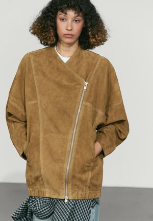 Donna con capelli ricci che indossa una giacca in camoscio color cuoio con una zip frontale asimmetrica e le mani nelle tasche, in piedi davanti a uno sfondo semplice.
