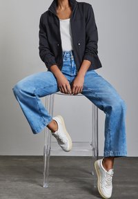 Henkilö istuu läpinäkyvällä tuolilla, päällään musta vetoketjullinen takki, valkoinen paita, vaaleansiniset leveälahkeiset farkut ja valkoiset tennarit.