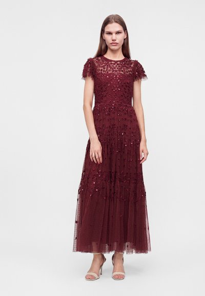 Jeune femme portant une robe longue en dentelle bordeaux avec broderies florales et manches courtes, debout devant un fond clair uni.