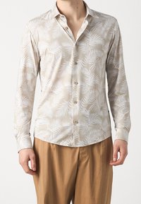 Helle beige langærmet skjorte med hvid palmebladsmønster; knapper foran og ved mansjetterne; blødt stof; buet kant. Bæres med brune bukser.