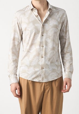Chemise beige clair à manches longues avec un motif de feuilles de palmier blanches ; boutons sur le devant et aux poignets ; tissu doux ; ourlet arrondi. Portée avec un pantalon beige.