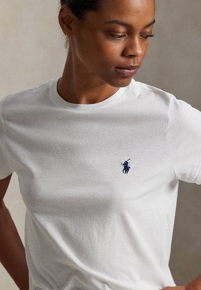 Kvinde iført en hvid t-shirt med korte ærmer og et lille mørkeblåt Polo-logo på brystet, som kigger ned og til siden.