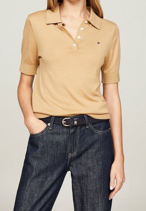 Vrouw draagt een beige poloshirt met korte mouwen, witte knopen en een klein logo, ingestopt in een donkerblauwe spijkerbroek met een zwarte riem.